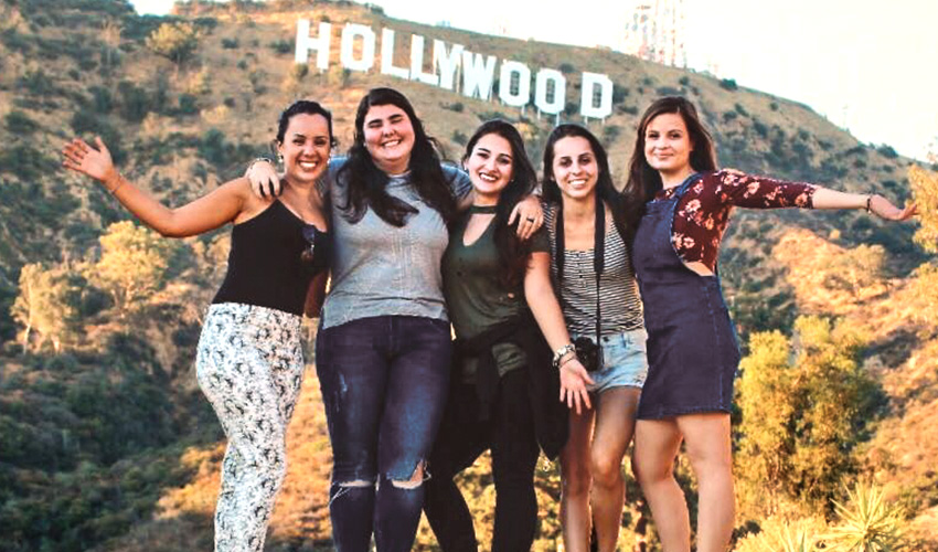 Five young women, students, smile together in front of the iconic Hollywood sign, capturing a memorable moment.