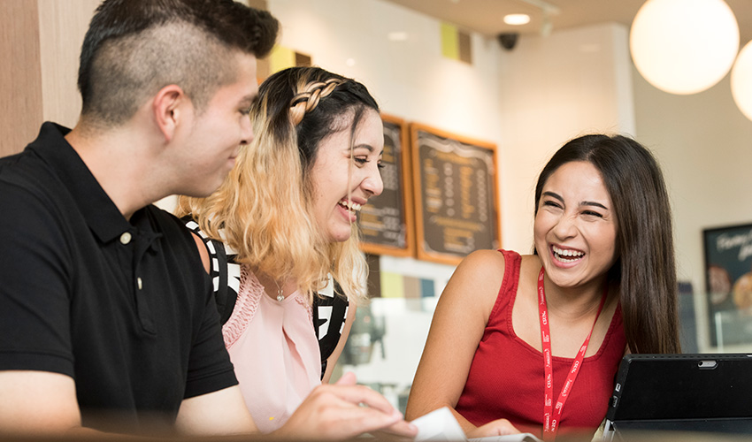 In a coffee shop, three students engage in discussion while working on a laptop at their table.