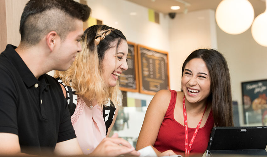 Three students gather at a coffee shop table, sharing ideas and working together on a laptop.