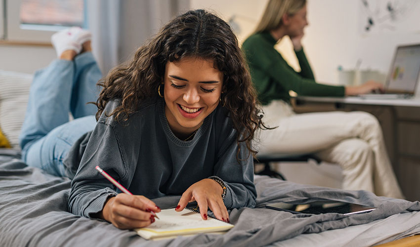 In her student housing room, a girl writes intently in a notebook while comfortably seated on her bed.