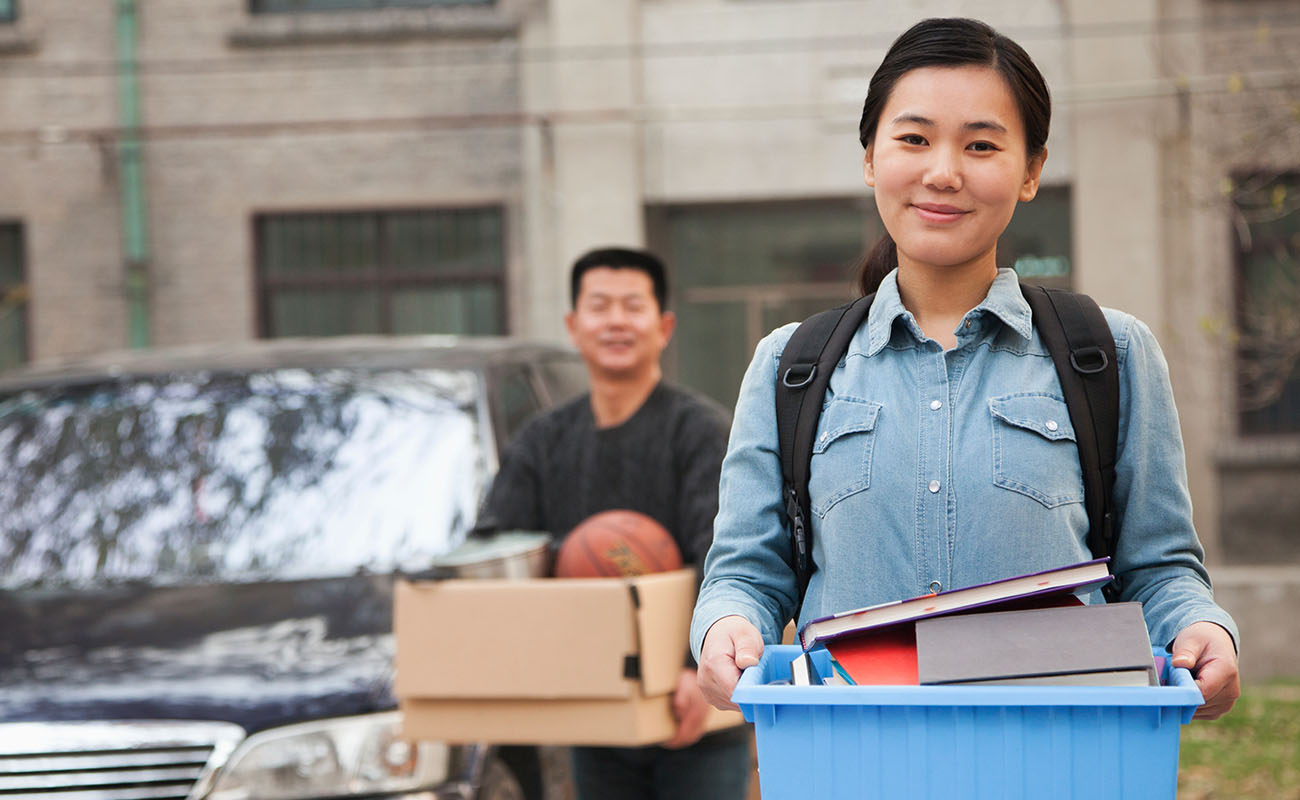 A woman stands in student housing, holding a blue container filled with books beside a parked car.