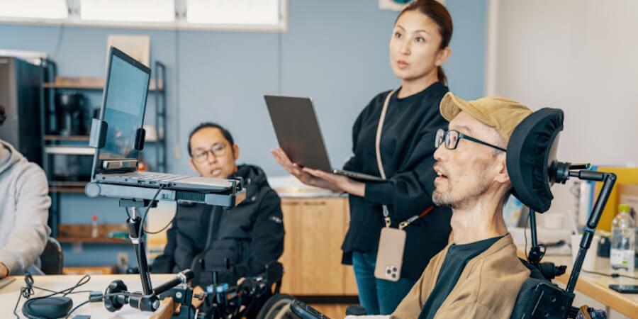 An assistive technology professional looks at computer equipment with two people who are in wheelchairs in an office.